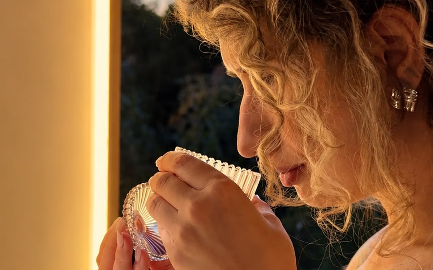 Invitată care testează un parfum, ținând în mâini un bol de sticlă cu esențe aromatice, în lumina caldă a standului